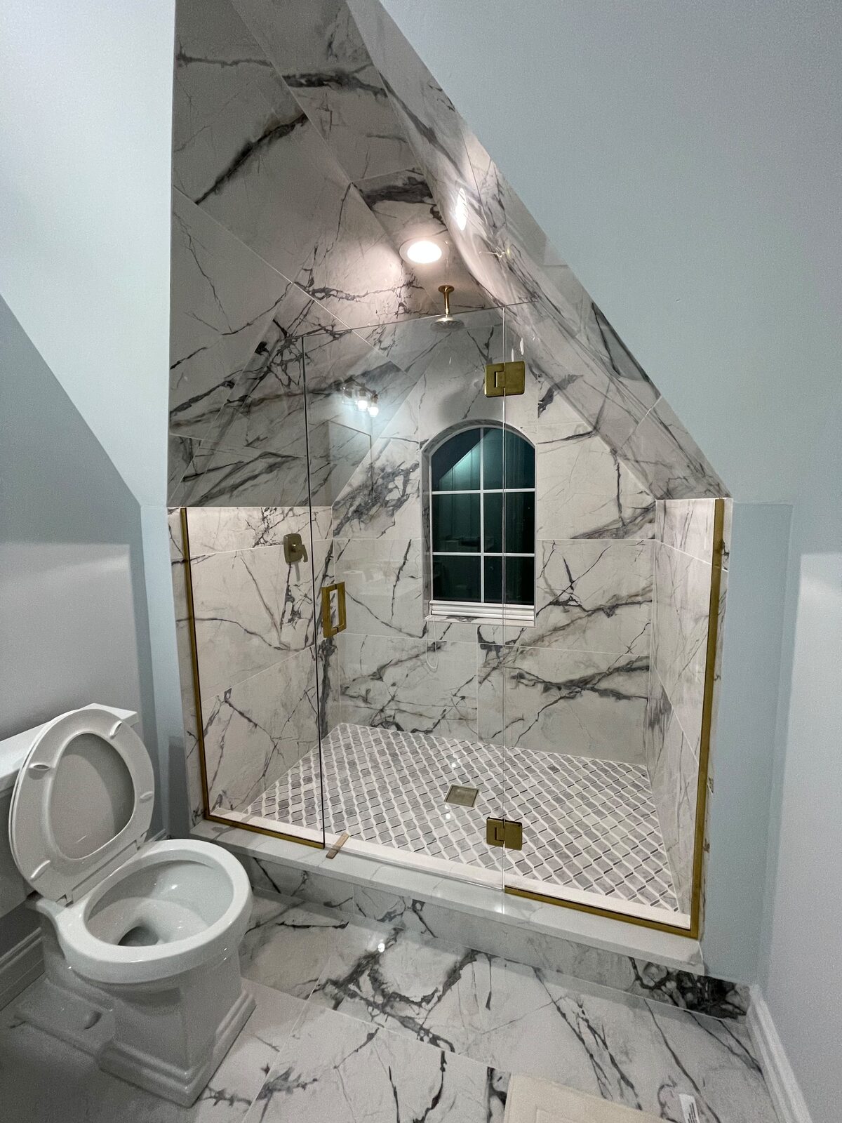Frameless glass shower under a dormer with arched window and gold trim